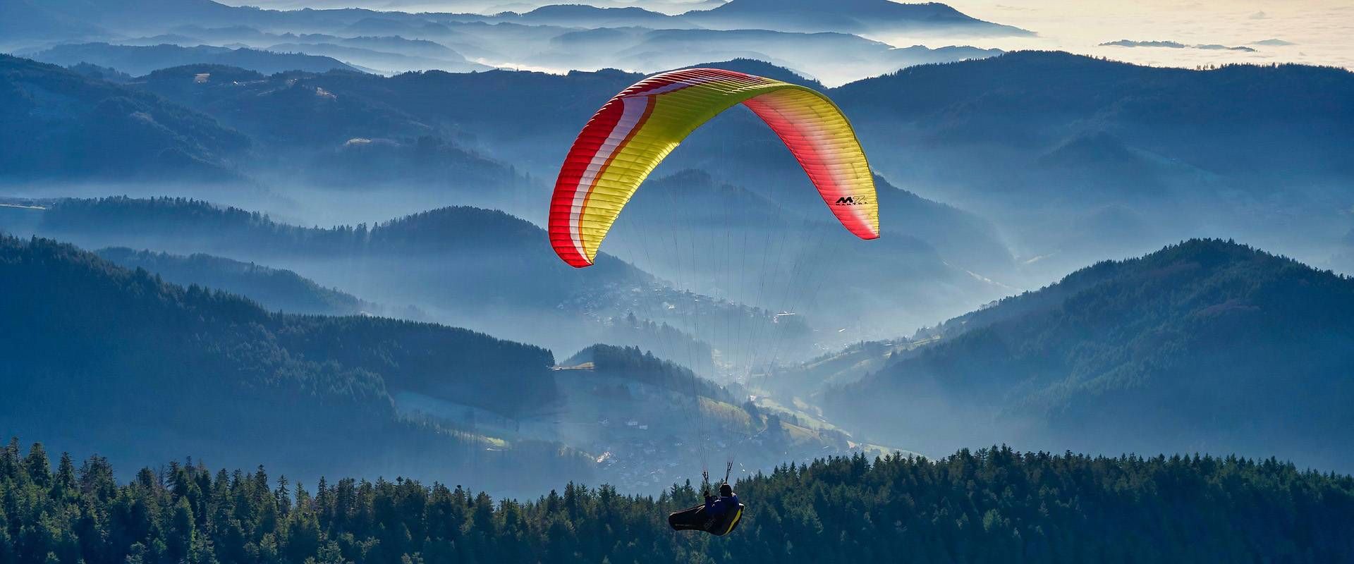 Ein Paraglider schwebt über einer bergigen Landschaft mit Wäldern und Nebelschichten im Tal.
