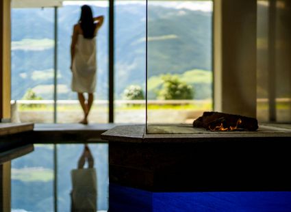 Eine Frau steht im Spa-Bereich am Fenster vor der Bergkulisse, daneben ein Becken mit Wasser und Feuerstelle.