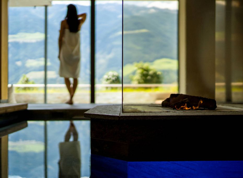 Eine Frau steht im Spa-Bereich am Fenster vor der Bergkulisse, daneben ein Becken mit Wasser und Feuerstelle.