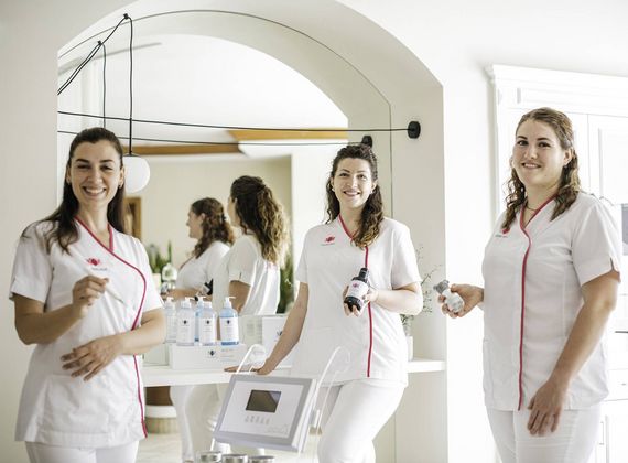 Drei Mitarbeiterinnen in weißen Uniformen stehen in einem hellen Behandlungsraum mit Spiegel und Pflegelotionen.