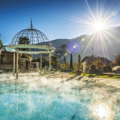 Ein Außenpool mit aufsteigendem Dampf, Pavillon und Sonnenlicht vor Bergkulisse im Preidlhof.