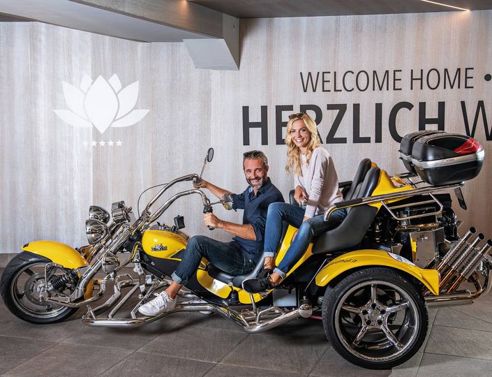 A smiling couple sits on a yellow three-wheeled motorcycle in front of a welcome wall.
