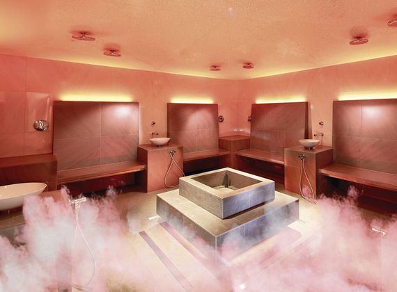A sauna relaxation room with benches, washbasins and a central stone feature surrounded by mist.