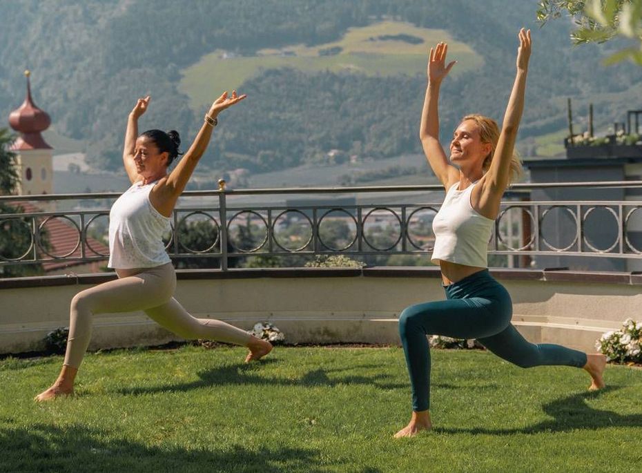 Zwei Frauen üben Yoga auf einer Rasenfläche mit Bergblick und Terrasse im Hintergrund.