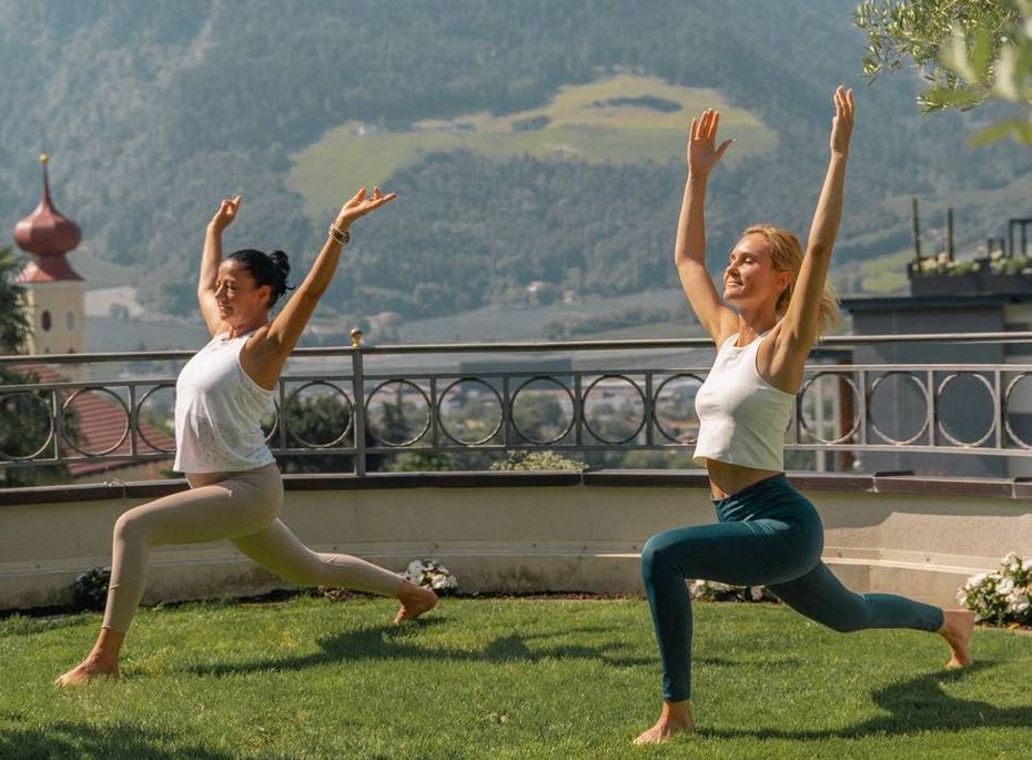 Zwei Frauen üben Yoga auf einer Rasenfläche mit Bergblick und Terrasse im Hintergrund.