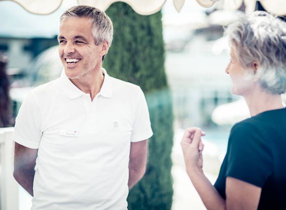 A smiling staff member in a white polo speaks with a woman in a spa setting.