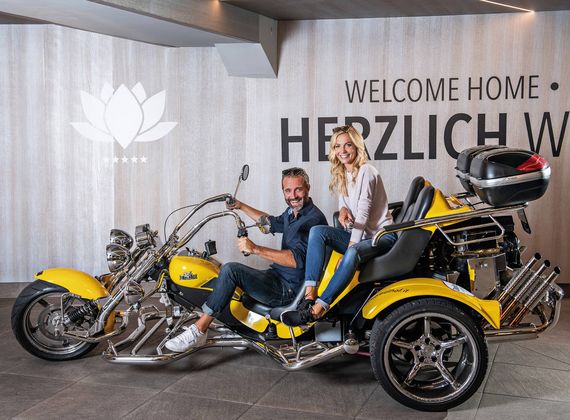 Ein Mann und eine Frau sitzen auf einem gelben Trike vor einer Wand mit der Aufschrift „Welcome home“.