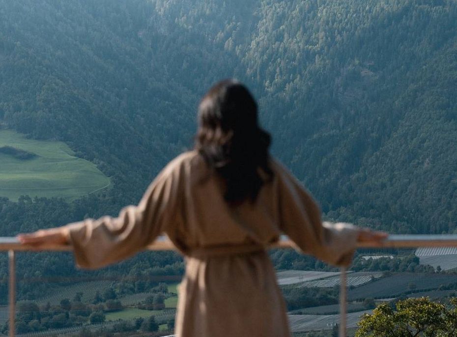 Wellnessausblick auf die Berge in Naturns Eine Frau im Bademantel steht auf einer Terrasse und blickt über das Tal und die Berge.
