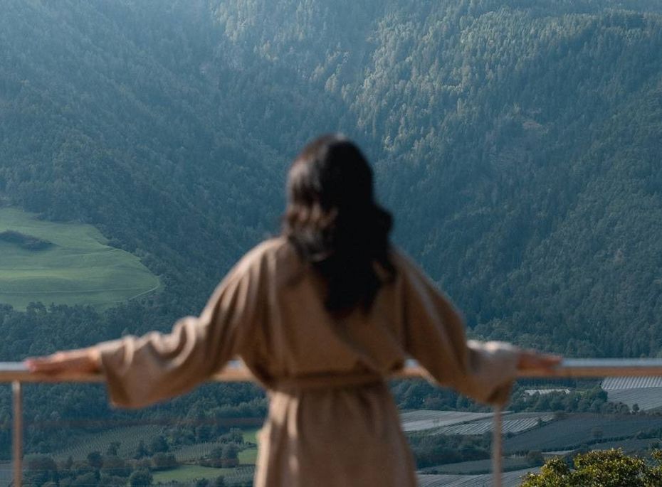 Wellnessausblick auf die Berge in Naturns Eine Frau im Bademantel steht auf einer Terrasse und blickt über das Tal und die Berge.