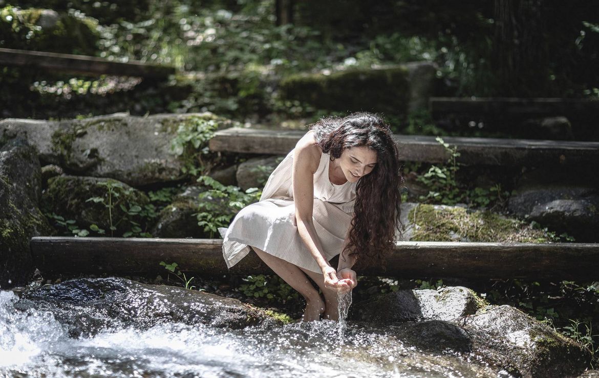 Eine Frau kniet an einem natürlichen Wasserlauf und lässt Wasser über ihre Hände fließen.