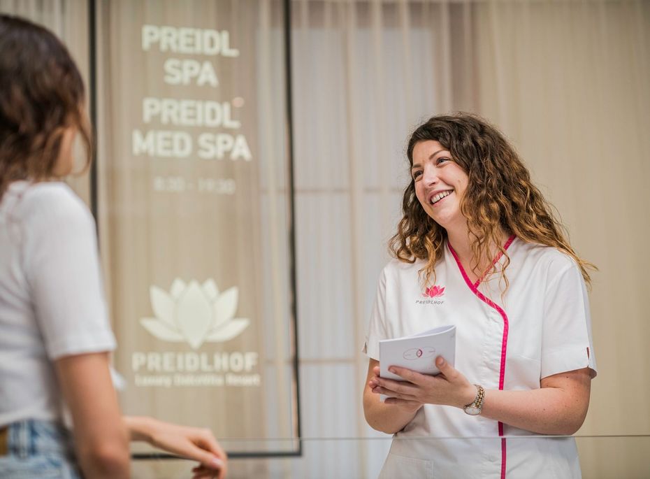 Preidlhof MED SPA Beratung am Empfang Eine Mitarbeiterin im hellen Preidlhof MED SPA spricht freundlich mit einer Besucherin am Empfang.